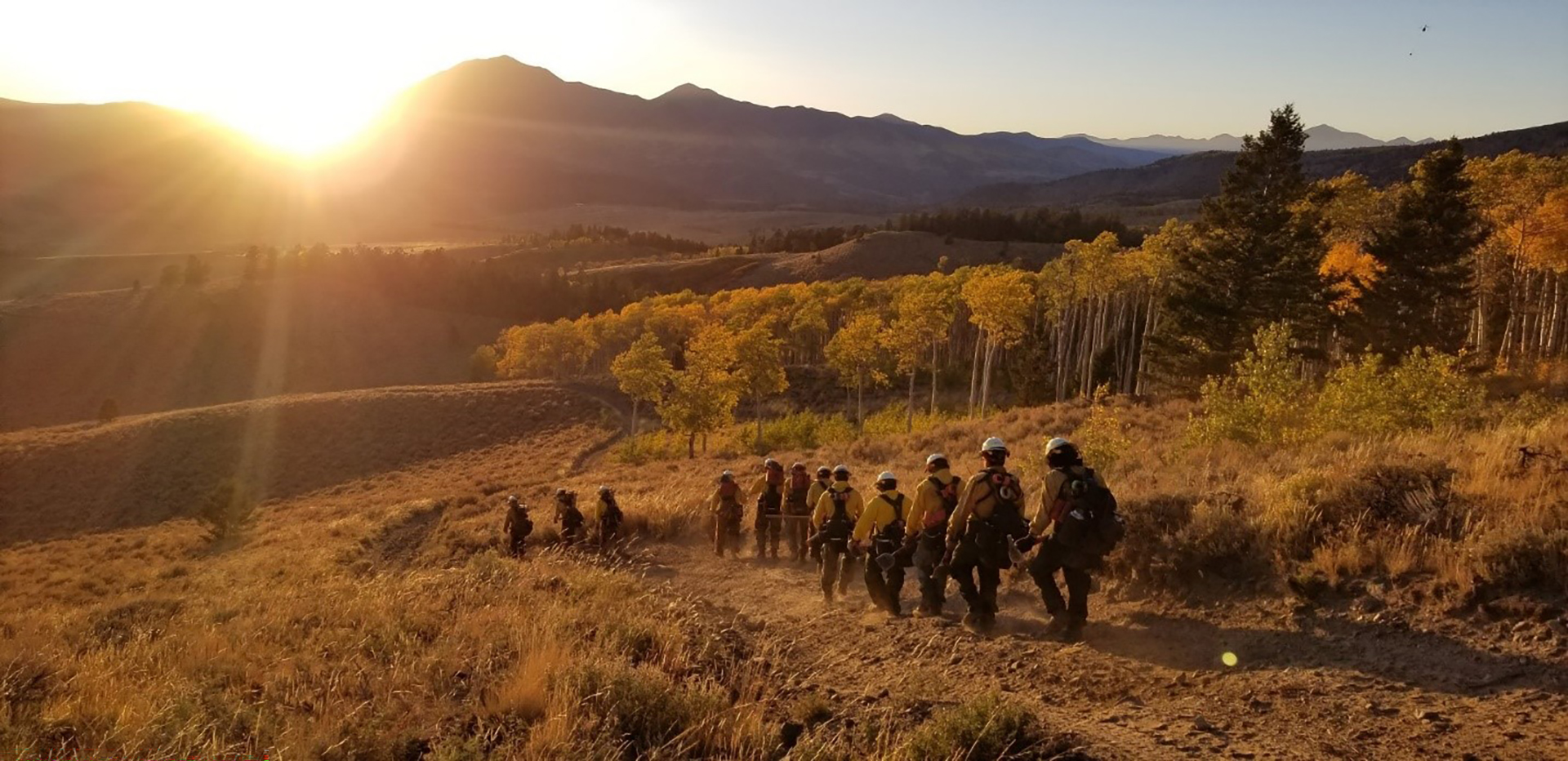 Crew of firefighters walk towards mountains at sunset. 