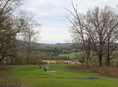A paved path with concrete pads leading to waysides signs lead out to a hill edge overlooking the Hudson River Valley with rolling hills in the background.