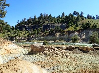 Rocky land scape next to white water river with construction vehicles in the grassy background.