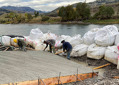 Three construction workers work on wooden boat ramp with bright blue river and green hills in the background. 