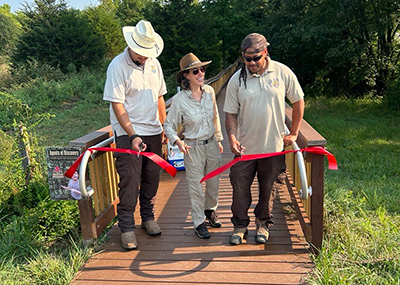 Staff at Patuxent Research Refuge cut red ribbon at the entrance of newly rehabilitated boardwalk surrounded by grass and trees.