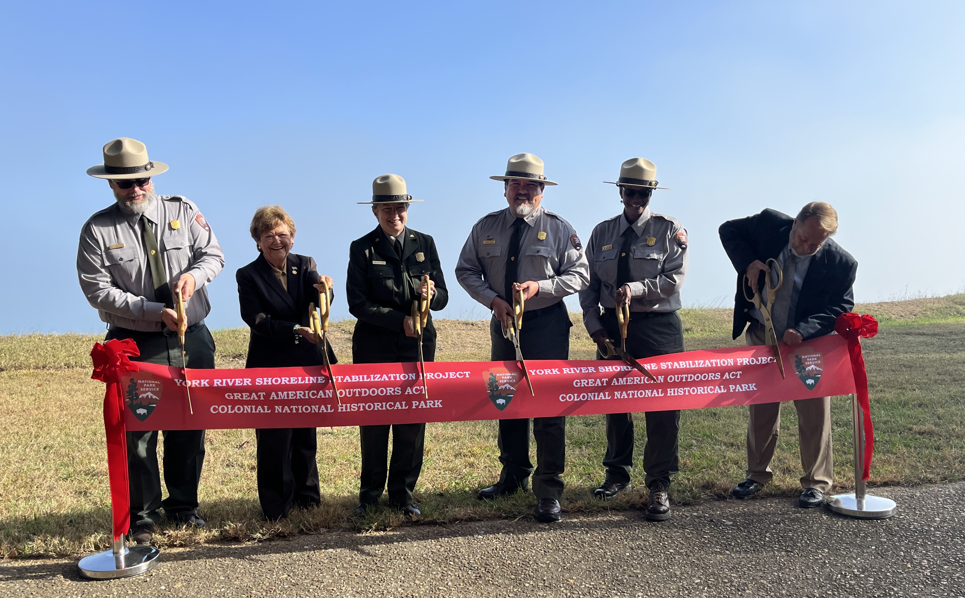 Six people, including four wearing National Park Service uniforms and two in business suits, pose with giant scissors to cut a red ribbon that says York River Shoreline Stabilization Project, Great American Outdoors Act, Colonial National Historical Park