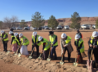 Group of people in yellow construction vests and hard hats use shovels to break ground on dirt plot.