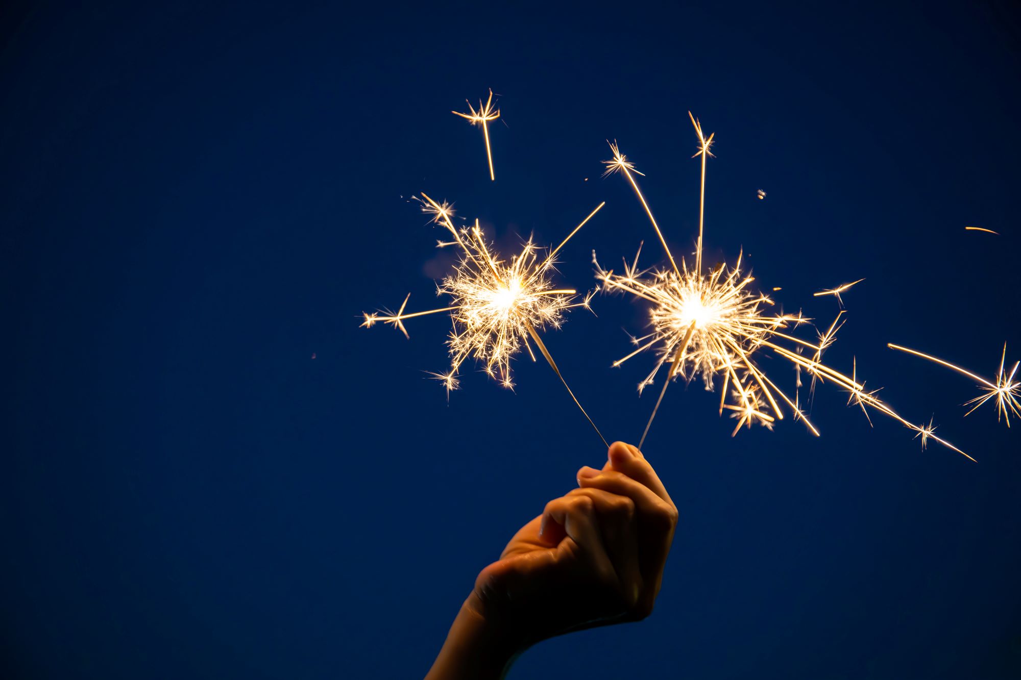 A hand holds two sparklers that are shooting sparks with a dark blue background.
