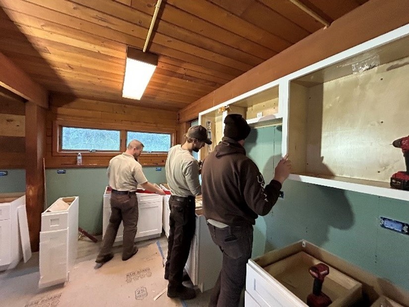 Three MAT members hang cabinets in a room with green walls and board ceiling.