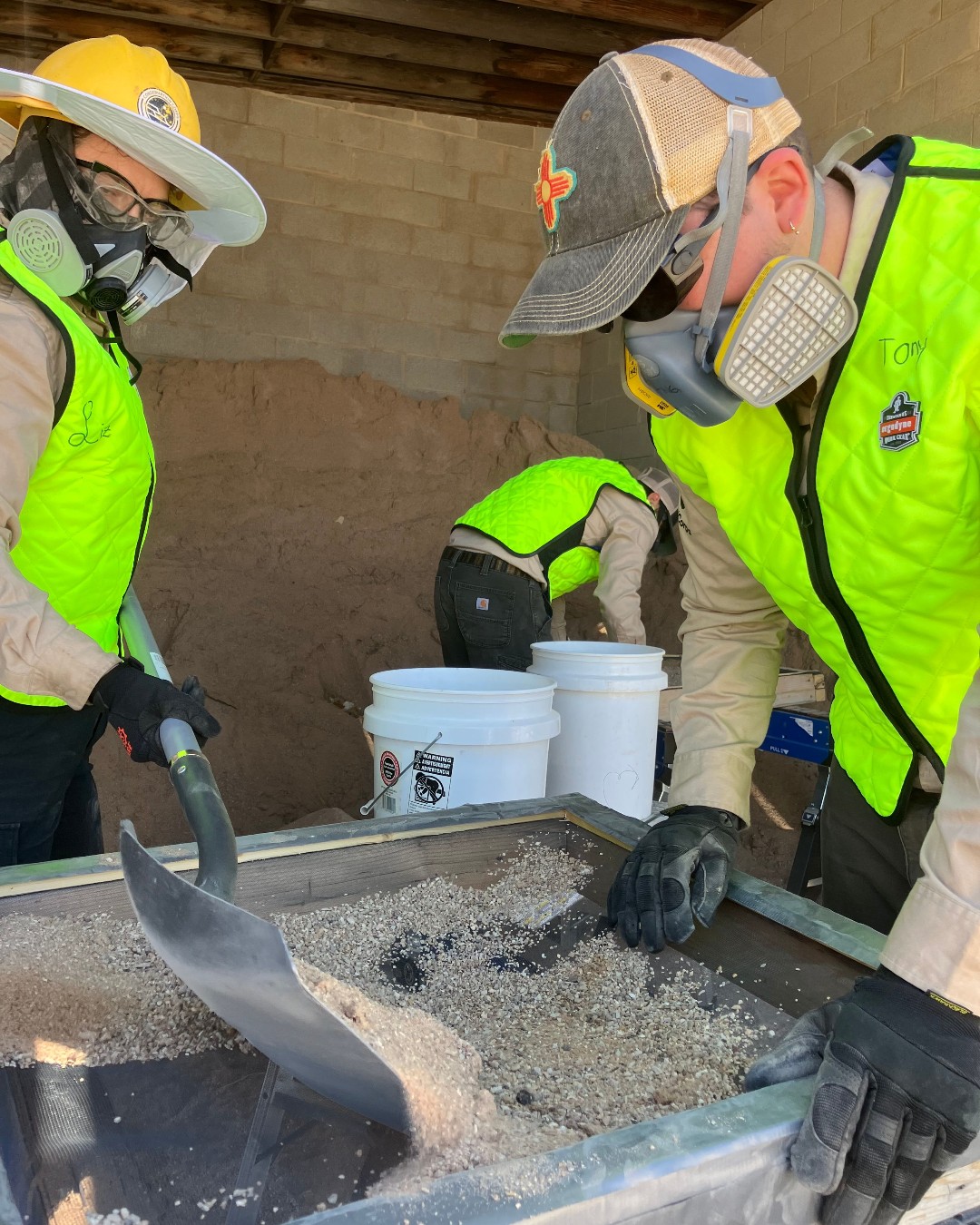 Three people wearing masks and yellow construction vests sift through sand in a metal tray.
