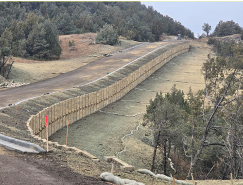 A dirt road in construction has a completed retaining wall and fresh hydroseeding; the road curves through a high desert pinyon juniper forest.