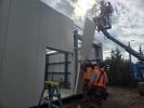 Men in orange vests and hard hats install gray wall panels to the side of a new building.