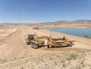 Construction vehicles sit on the dirt perimeter of large body of water.