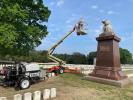 One person uses construction equipment to elevate above a statue of a weeping solder; in the background rows of aligned white headstones of the cemetery are seen.