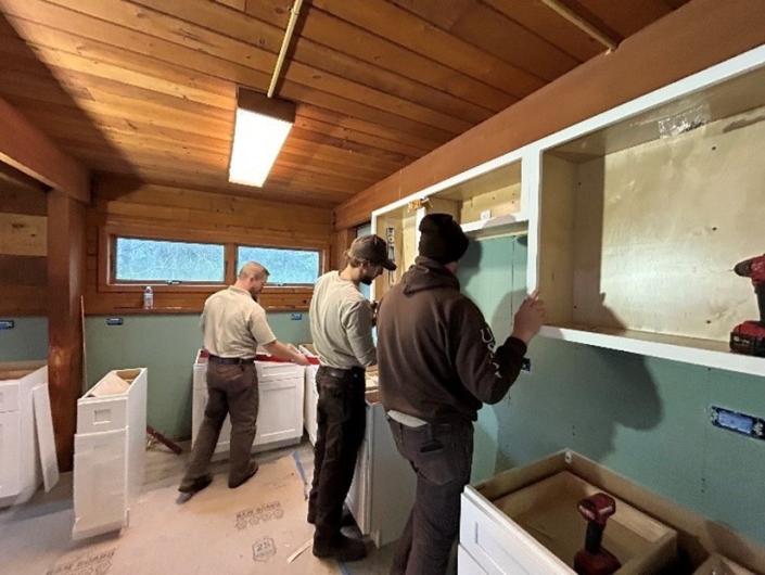 Three MAT members hang cabinets in a room with green walls and board ceiling.