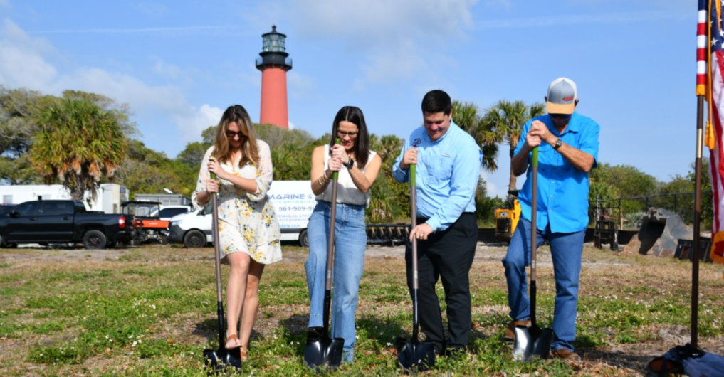  Four people use shovels to break ground on grass with a red lighthouse rising above the trees in the background.