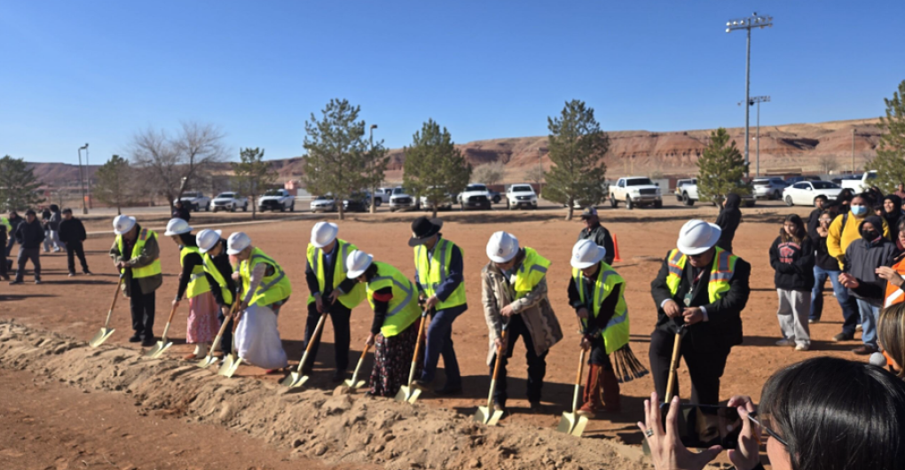 Group of people in yellow construction vests and hard hats use shovels to break ground on dirt plot.