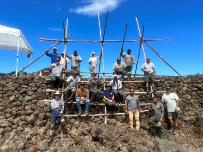 A group of people sit on a wooden ladder system in front of a wall of rocks.