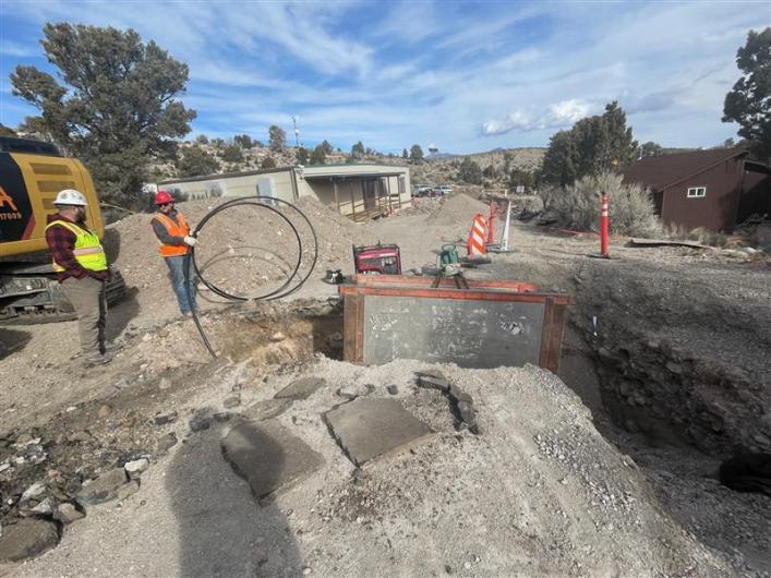 Men in construction vests and hard hats stand next to a large hole with water system components in it.