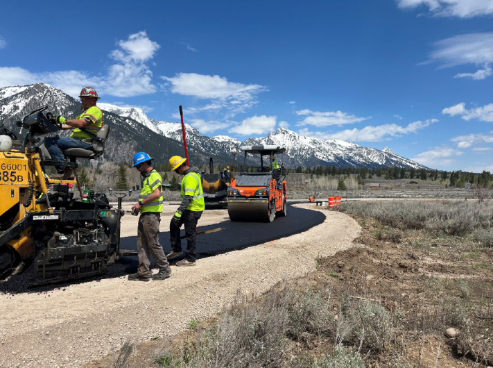 Construction men in yellow vests lay asphalt on road with snow-capped mountains in the background.