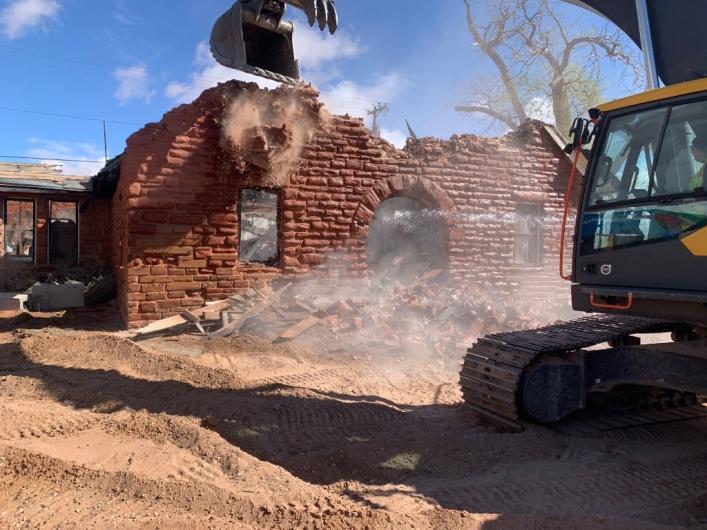Yellow construction vehicle demolishes red brick building in construction area.