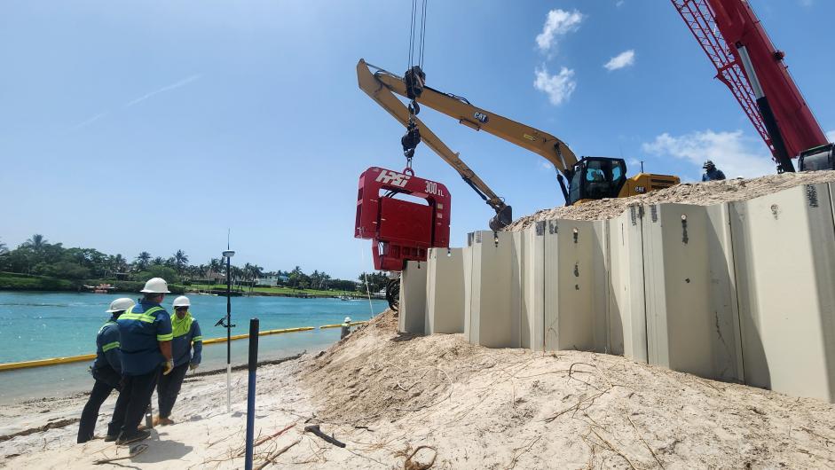 Three men in hard hats stand next to concrete walls blocking a large sand pile and construction vehicles.