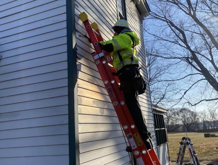 Man wearing a yellow vest and hard hat stands on a ladder propped against the side of a white house.​