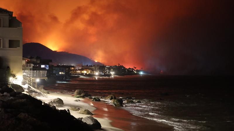 A wildfire near Los Angeles burns at night. The beach is in the foreground, with houses lining the coast as a red glow and smoke rise in the distance. CAL FIRE image.