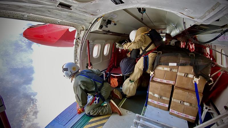 The cylindrical interior of an airplane is filled with strapped down boxes and a smokejumper wearing bulky tan gear with supplies in black bags strapped to his harness and secured in large pockets. One person in a helmet kneels by the open side door of the plane, leaning forward to look down at smoke rising from a wildfire below.