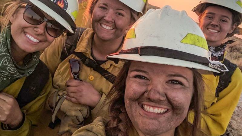 Four women with soot-smudged faces pose for a selfie, smiling at the camera. They are wearing wildland firefighter yellow shirts, white helmets with yellow visibility strips, and the black straps of packs on their shoulders. One holds a pair of sunglasses. Two others have bandanas around their necks. Reddish light illuminates the two in the back from the side. Behind them, a hint of the landscape is just visible with dry shrubs bordering hard-packed ground. The photo is taken at an angle so that the horizon