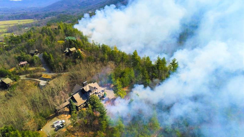 A large house with two trucks parked outside is seen from above. A handful of other, smaller homes are scattered through the surrounding forested area, connected by narrow, winding roads. White smoke rises in a wedge formation from the trees to the right of the homes, obscuring the forest from view. To the left, the forested hillside descends toward a grassy plain, with more forested mountains visible in the distance.
