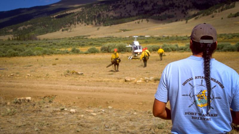 A firefighter with a long braid and wearing a Fort Washakie Helitack T-shirt stands in the foreground, his back to the camera. He’s watching firefighters approach a helicopter on the other side of a dirt track on an expanse of dusty ground. In the background, mountains with a patchwork of trees and dry grass are studded with the shadows of passing white clouds.