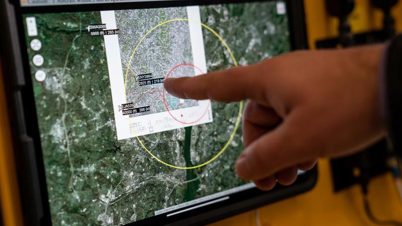  A hand extends toward a tablet screen mounted on a yellow panel, a finger pointing toward highlighted text and a red circle near the center of the screen. The screen displays a detailed topographical map with overlaid concentric circles and three small airplane icons with associated text. A portion of the map is magnified in an overlaid callout box.