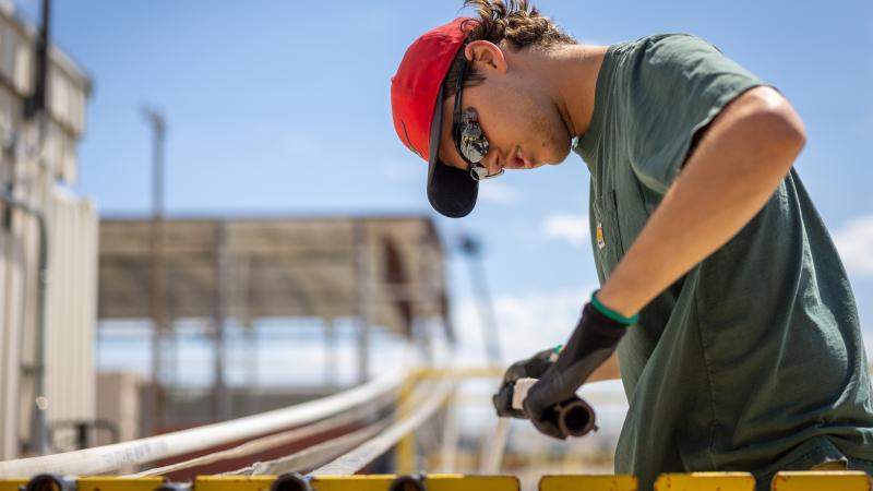 A man in a T-shirt, baseball hat, sunglasses, and protective gloves manipulates a nozzle in his hands while leaning forward for a closer look. He’s standing outside under a bright blue sky on an expanse of concrete where a large, yellow, metal stand is holding lengths of hose stretched between each end. To the side of his workspace, the corner of an industrial building is just visible. Behind it is tall roof over an open space.