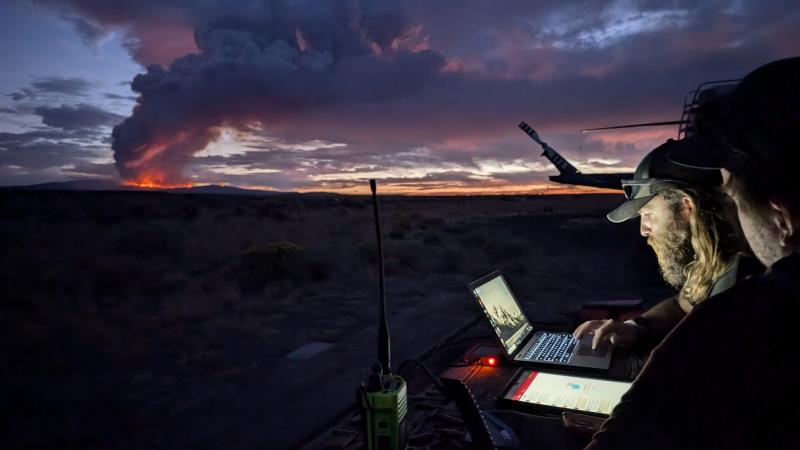 Two people work on a laptop and a tablet at dusk, standing at a makeshift table set up in a field of sagebrush. A radio and other equipment clutter the table. Their faces are illuminated by the glow of the screens. The tail of a helicopter is visible behind them. An orange-red fire glows on the mountains in the distance, a plume of smoke rising into the sky and merging with the scattered clouds that are lit by the last light of the sunset.