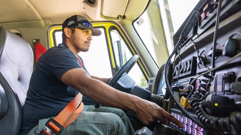 A wildland firefighter sits inside the cab of a wildland fire engine. He is pointing his finger at one of the equipment buttons as he operates the engine.