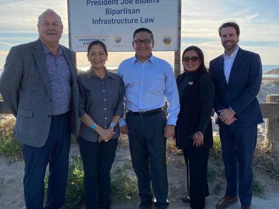 Secretary Haaland and four others standing near beach in front of Investing in America sign.
