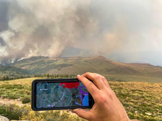 A cell phone displaying a real-time map of a wildfire is held up in front of hills where smoke from the fire can be seen rising into the sky.