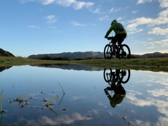 A rider wearing a helmet rides an e-bike past a small pond reflecting the clouds and sky