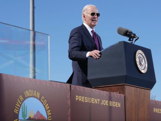 President Biden stands behind a podium in a dark suit and purple tie and delivers remarks