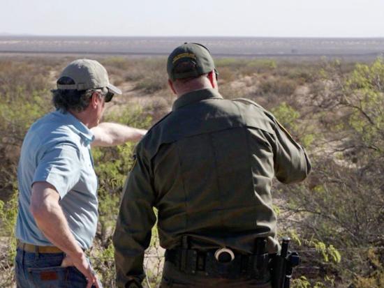 Secretary Burgum in a ballcap, jeans and blue shirt points across an open desert plain next to a green uniformed Border Patrol agent