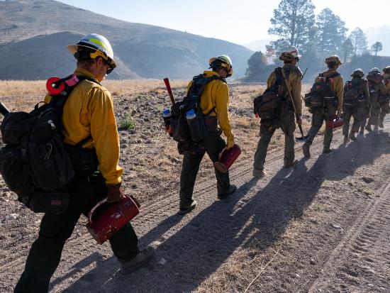 Wildland firefighters hike in a line wearing wildland firefighting gear and carrying tools. 