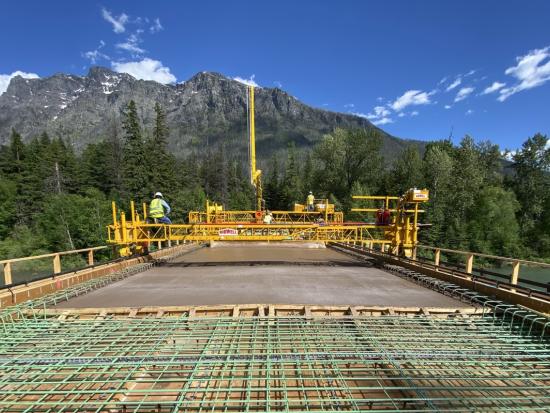 Against a backdrop of mountains and pine trees, a bridge is being constructed.