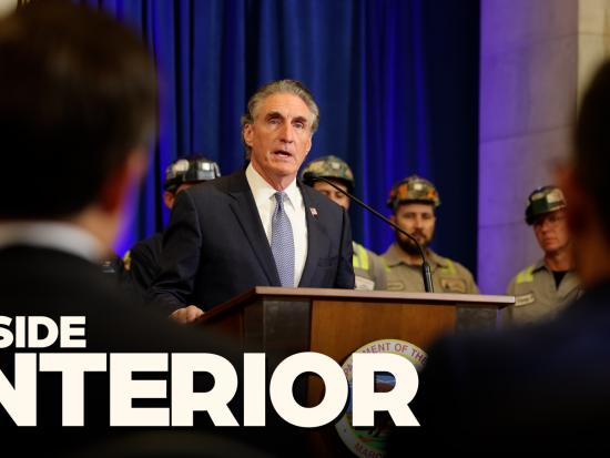Secretary Burgum stands behind a wooden and metal podium in front of guests and flags and delivers remarks to a crowd