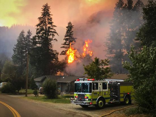 A ranch house sits on a quiet one-lane road, backing up to a forested hillside. A fire engine is parked in the driveway, a hose snaking along the ground beside it. Immediately behind the house, yellow-orange flames blaze through several evergreen trees. Smoke obscures the forested hills beyond the neighborhood.