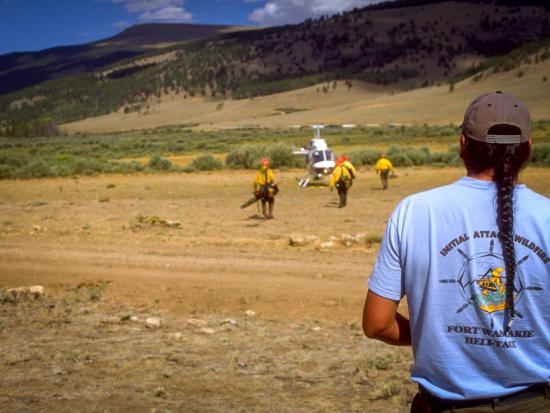 A firefighter with a long braid and wearing a Fort Washakie Helitack T-shirt stands in the foreground, his back to the camera. He’s watching firefighters approach a helicopter on the other side of a dirt track on an expanse of dusty ground. In the background, mountains with a patchwork of trees and dry grass are studded with the shadows of passing white clouds.