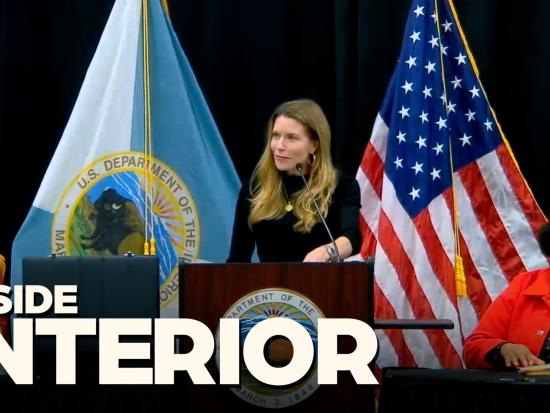 Leslie Beyer stands at a podium in front of the American Flag and the Interior Department Flag