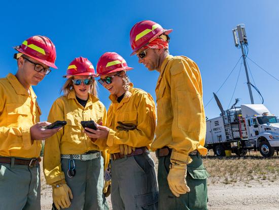 Four wildland firefighters cluster together during the Vader Fire looking down at two mobile devices and smiling. Behind them, a FirstNet satellite cell truck is parked on a dry field with a large antennae deployed.
