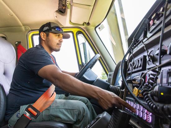 A wildland firefighter sits inside the cab of a wildland fire engine. He is pointing his finger at one of the equipment buttons as he operates the engine.
