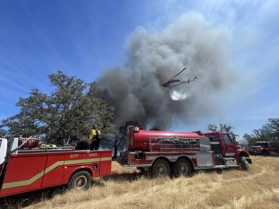 Two red firefighting vehicles are parked in a field of dry grass as smoke rises into a blue sky from a wildfire in the distance. A wildland firefighter stands on one of the vehicles, which is a small wildland fire engine, as he looks towards the rising smoke. A helicopter flies overhead, dropping water from a long hose attached to its belly.