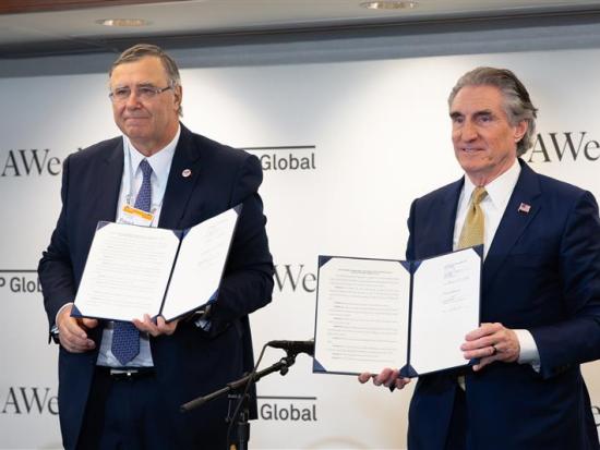 Two men in suits holding signed documents on a stage. 