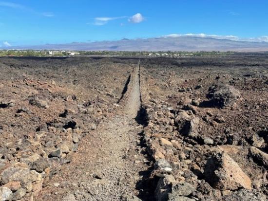A worn trail is cut through a rough gray and red lava flow, extending in a straight line to the horizon. A low mountain is in the background with blue sky above.