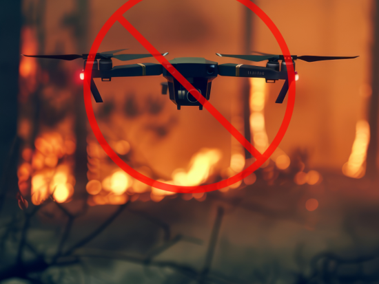 A drone hovers just above the ground, a wildfire burning behind it. A red circle with a line through it is shown over the drone.