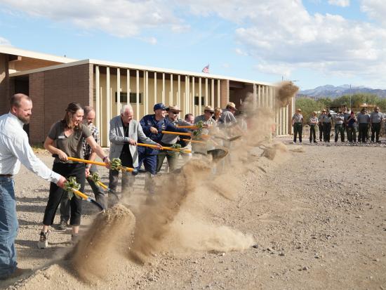 A line of people holding shovels with yellow handles throw a cloud of brown dirt while people watch.